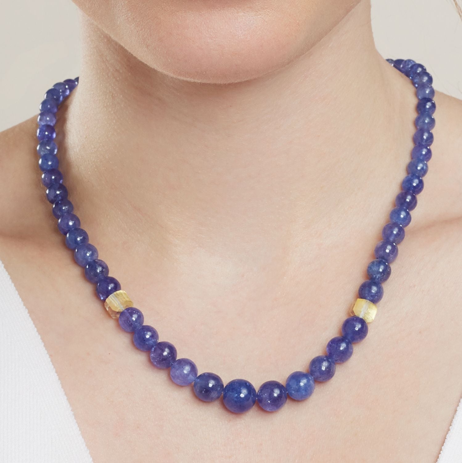 Ausschnitt des Halses einer Frau; sie trägt ein Collier aus direkt aneinander gereihten, blauen, runden Steinen; vom Stein in der Mitte aus jeweils 5 Steine rechts und links ist ein heller Stein eingefügt; die Steine werden zur Mitte hin größer;