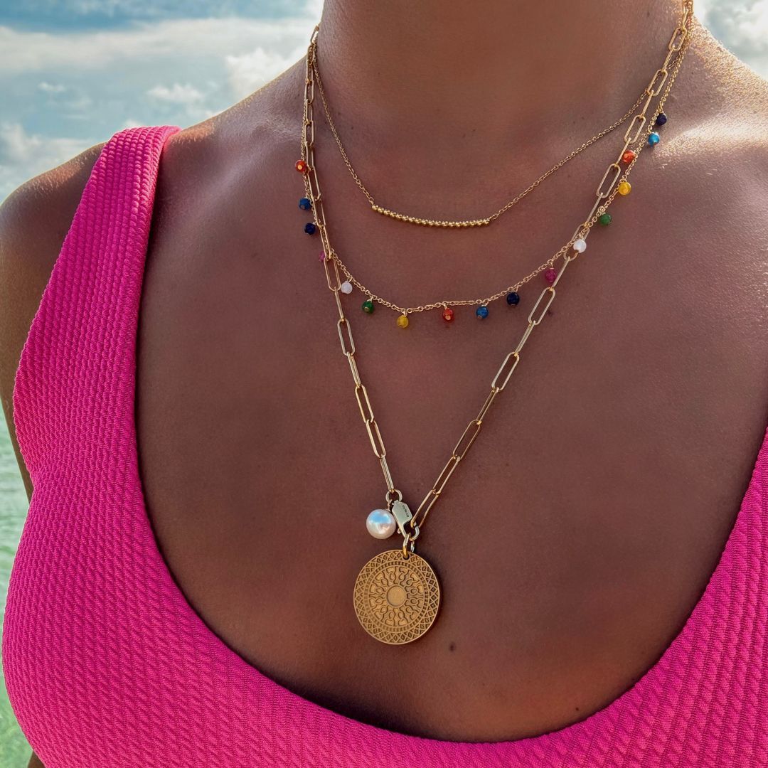 Halsausschnitt einer Frau; sie trägt eine goldene Gliederkette mit großen, feinen Gliedern, daran eine weiße Perle und ein goldener, runder Anhänger mit Musterung; eine goldene Gliederkette mit 17 bunten Steinen, die jeweils in kurzem Abstand an der Kette hängen; eine feine goldene Gliederkette; in der Mitte 18 kleine goldene Kugeln;