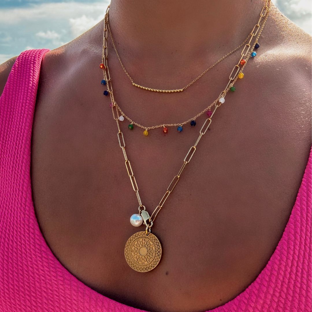 Halsausschnitt einer Frau; sie trägt eine goldene Gliederkette mit großen, feinen Gliedern, daran eine weiße Perle und ein goldener, runder Anhänger mit Musterung; eine goldene Gliederkette mit 17 bunten Steinen, die jeweils in kurzem Abstand an der Kette hängen; eine feine goldene Gliederkette; in der Mitte 18 kleine goldene Kugeln;