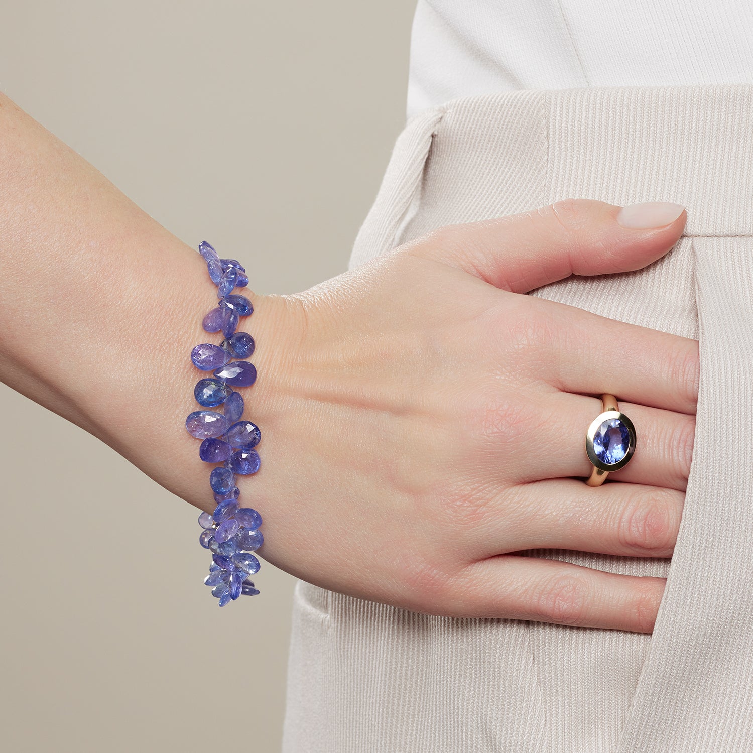 Ausschnitt einer Hand, die halb in einer Hosentasche steckt; am Arm ist ein Armband aus jeweils rechts und links direkt aneinander gereihten, blauen, tropfenförmigen Steinen; am Mittelfinger ist ein Ring aus Gelbgold mit einem in Gelbgold quer gefassten, blauen, ovalen Stein;