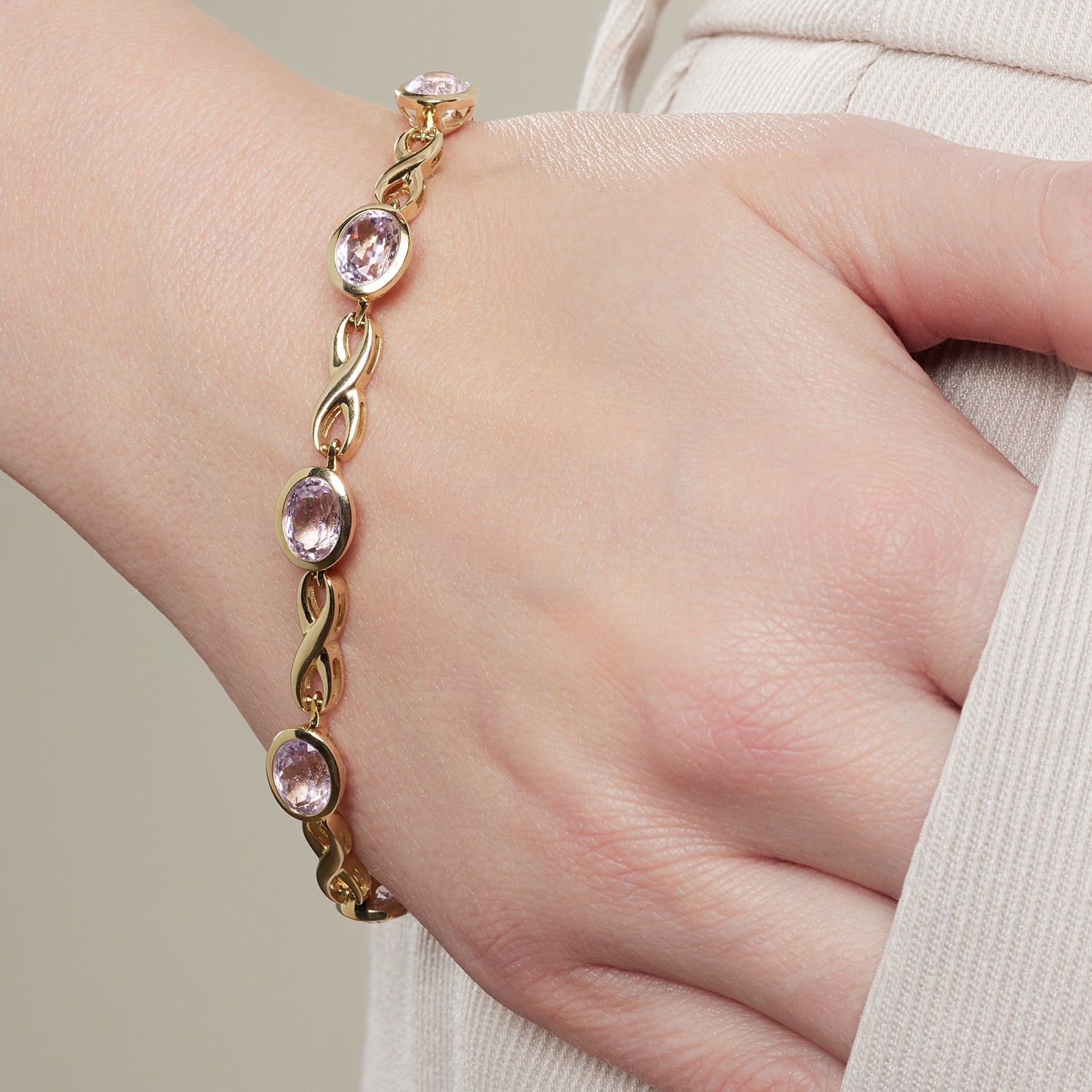 Ausschnitt einer Hand, die in einer Hosentasche steckt; an der Hand ein goldenes Armband mit eingefassten, helllila, ovalen Steinen; die Verbindung zwischen den einzelnen Steinen ähnelt einer liegenden Acht;