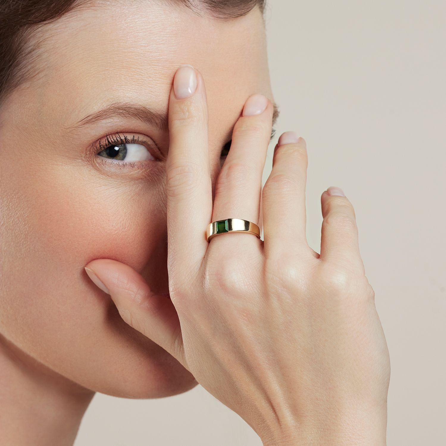 Ausschnitt des Gesichts einer Frau; sie hält ihre Hand davor und bedeckt ein Auge; am Mittelfinger trägt sie einen Band-Ring in Gelbgold, eingelegt ein viereckiger, dunkelgrüner Stein;