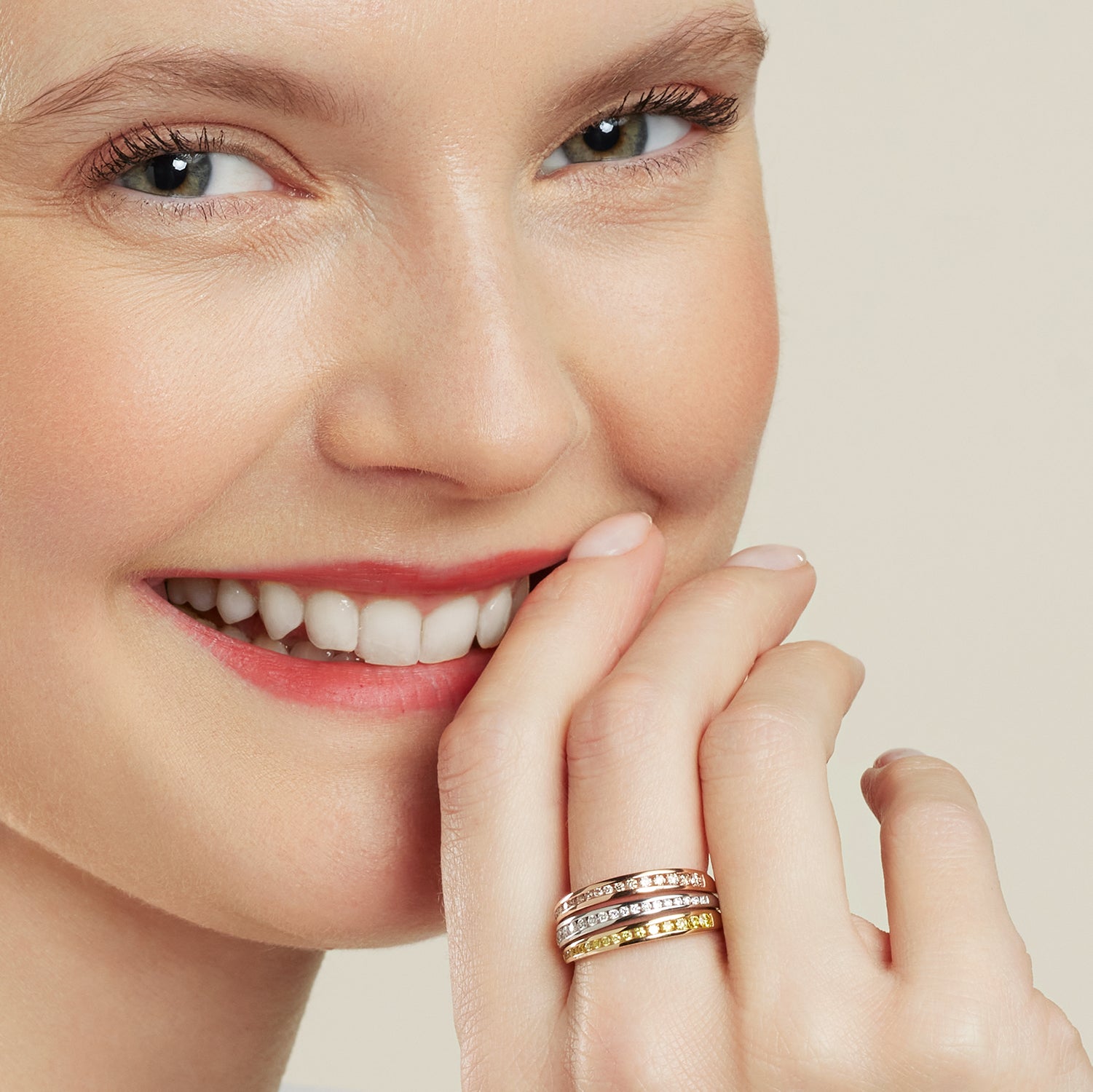 Gesicht einer lachenden Frau, die ihre Hand an den Mund hält; sie trägt am Mittelfinger einen Rivière-Ring mit einzelnen Band-Schienen aus Weiß-, Rosé- und Gelbgold; in jeder Band-Schien sind 8 Brillanten direkt aneinander eingefasst;