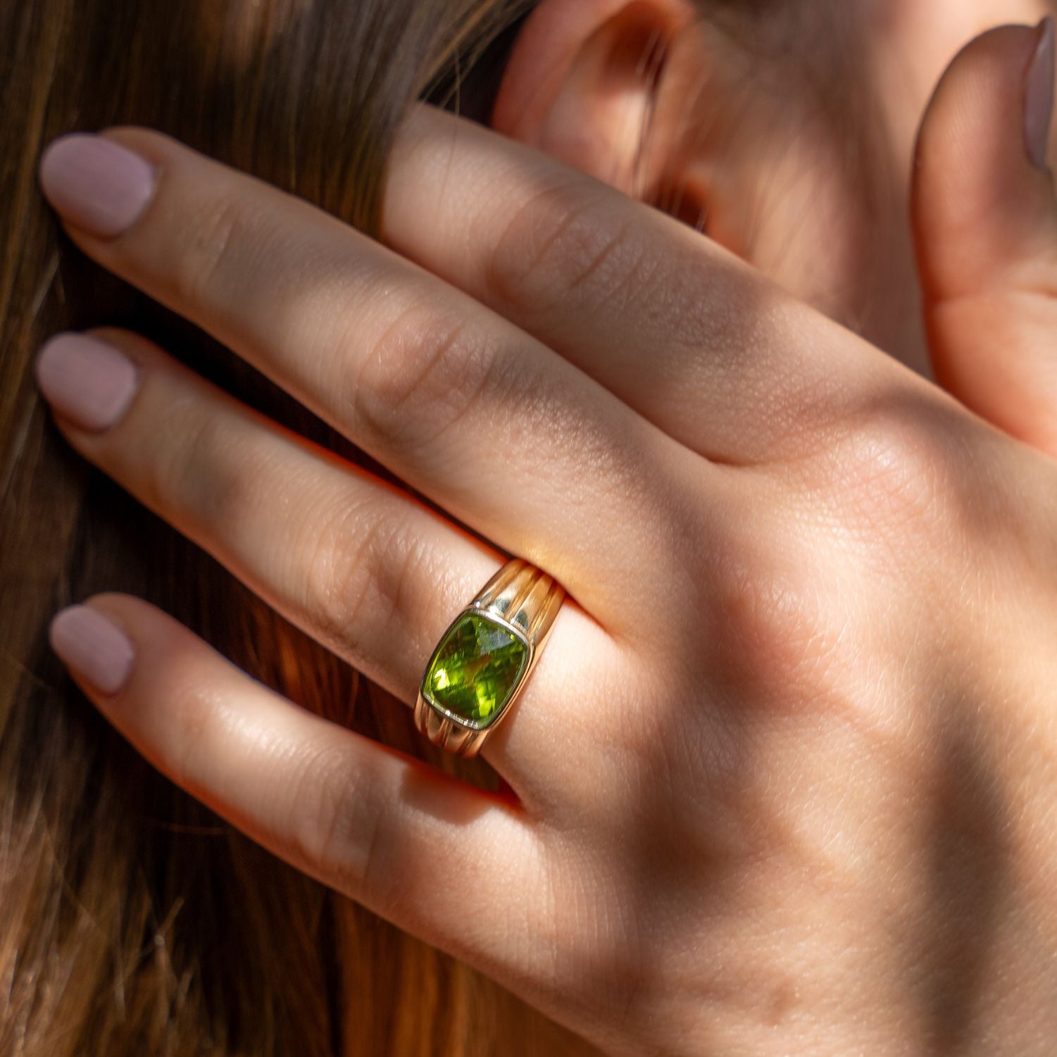 Ausschnitt einer Frauenhand, die an das Ohr fasst; am Ringfinger ist ein breiter, gerillter Band-Ring in Gelbgold mit einem eingefassten, viereckigen, grünen Stein;