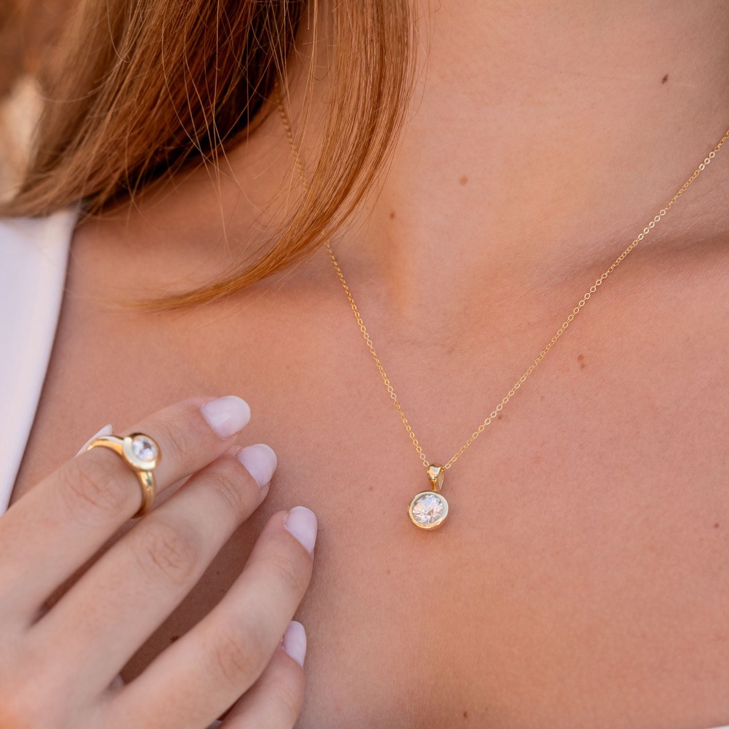 Ausschnitt des Halses und einer Hand einer Frau; sie trägt eine goldene Gliederkette mit einem Clip-Anhänger aus Gelbgold mit einem in Gelbgold eingefassten, runden, weißen Zirkon; am Zeigefinger steckt ein Band-Ring in Gelbgold mit einem in Gelbgold gefassten, runden, weißen Zirkon;