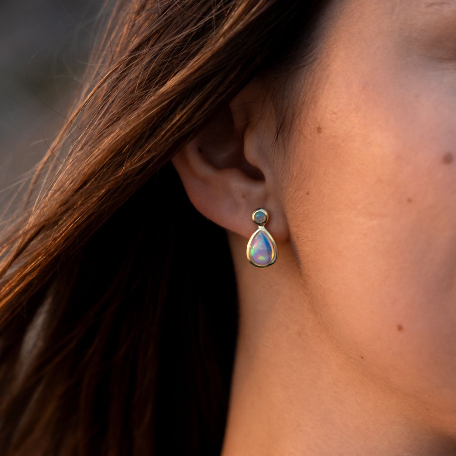 Ausschnitt des linken Ohres einer Frau mit langen dunklen Haaren; sie trägt einen Ohrstecker mit einem kleinen runden, bläulich schimmernden, silbervergoldet eingefassten Opal oben und einem tropfenförmigen größeren Opal unten;