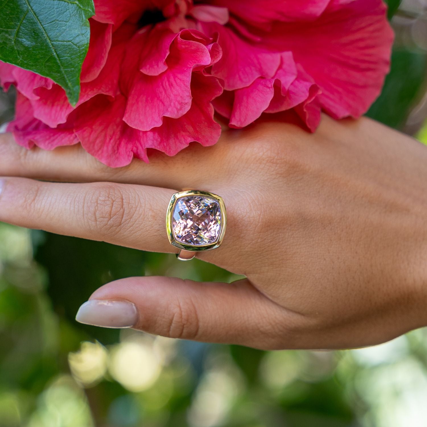 Eine Hand an einer Blume; am Ringfinger steckt ein goldener Ring mit einem golden eingefassten, viereckigen, helllila Stein;