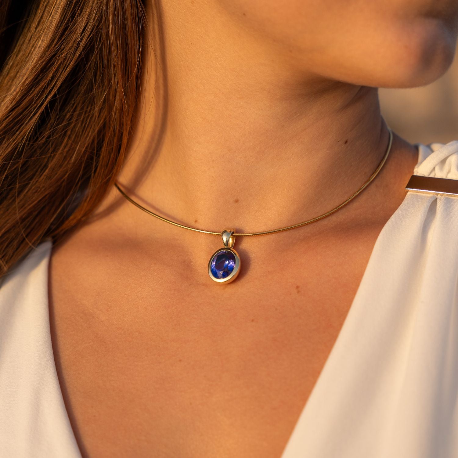 Ausschnitt des Halses einer Frau; sie trägt eine Schlangenkette mit einem Clip-Anhänger in Gelbgold mit einem in Gelbgold gefassten, runden, blauen Stein;