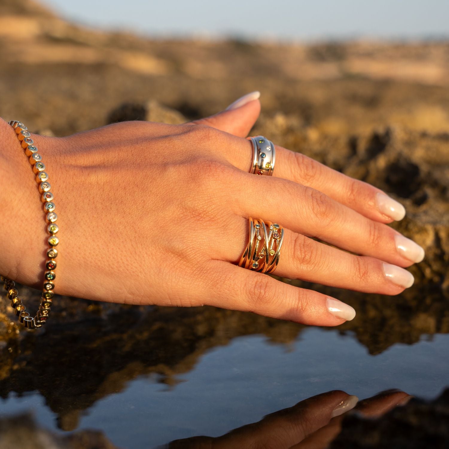 Die Hand einer Frau; im Hintergrund Wasser und Steine;
Sie trägt am Ringfinger einen breiten Ring in Gelbgold mit ineinander verschlungenen Ring-Schienen; diese sind besetzt mit in Gelbgold eingefassten bunten Brillanten; am Zeigefinger trägt sie einen breiten Band-Ring in Gelbgold mit vereinzelten bunten Brillanten; am Handgelenk ist ein Armband aus Gelbgold mit direkt aneinander gereihten, einzeln eingefassten bunten Brillanten in Gelbgold;