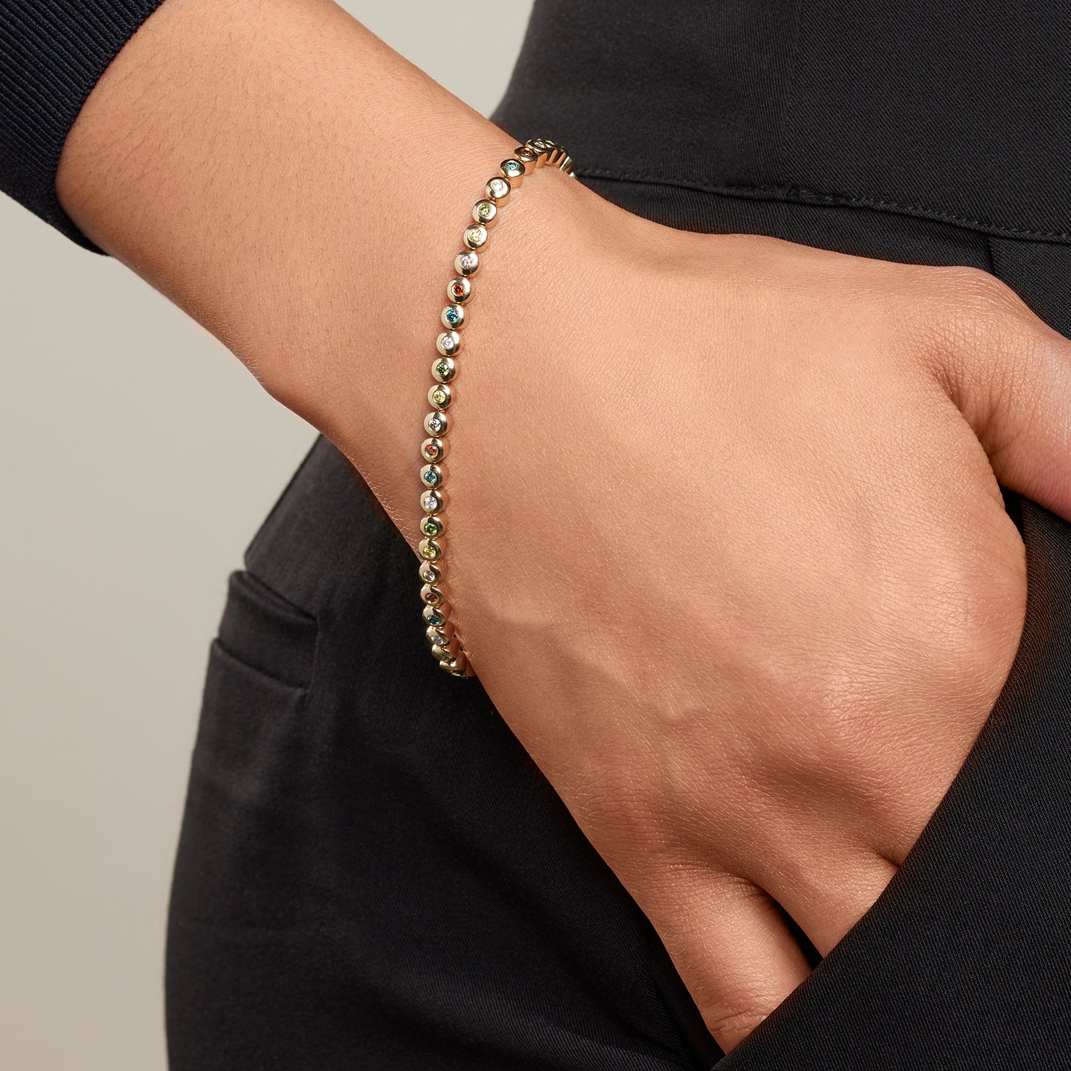 Die Hand einer Frau steckt halb in einer Hosentasche. Am Arm trägt sie ein Armband aus Gelbgold mit direkt aneinander gereihten, einzeln eingefassten Brillanten in Gelbgold in den Farben rot, blau, gelb, grün, weiß;
