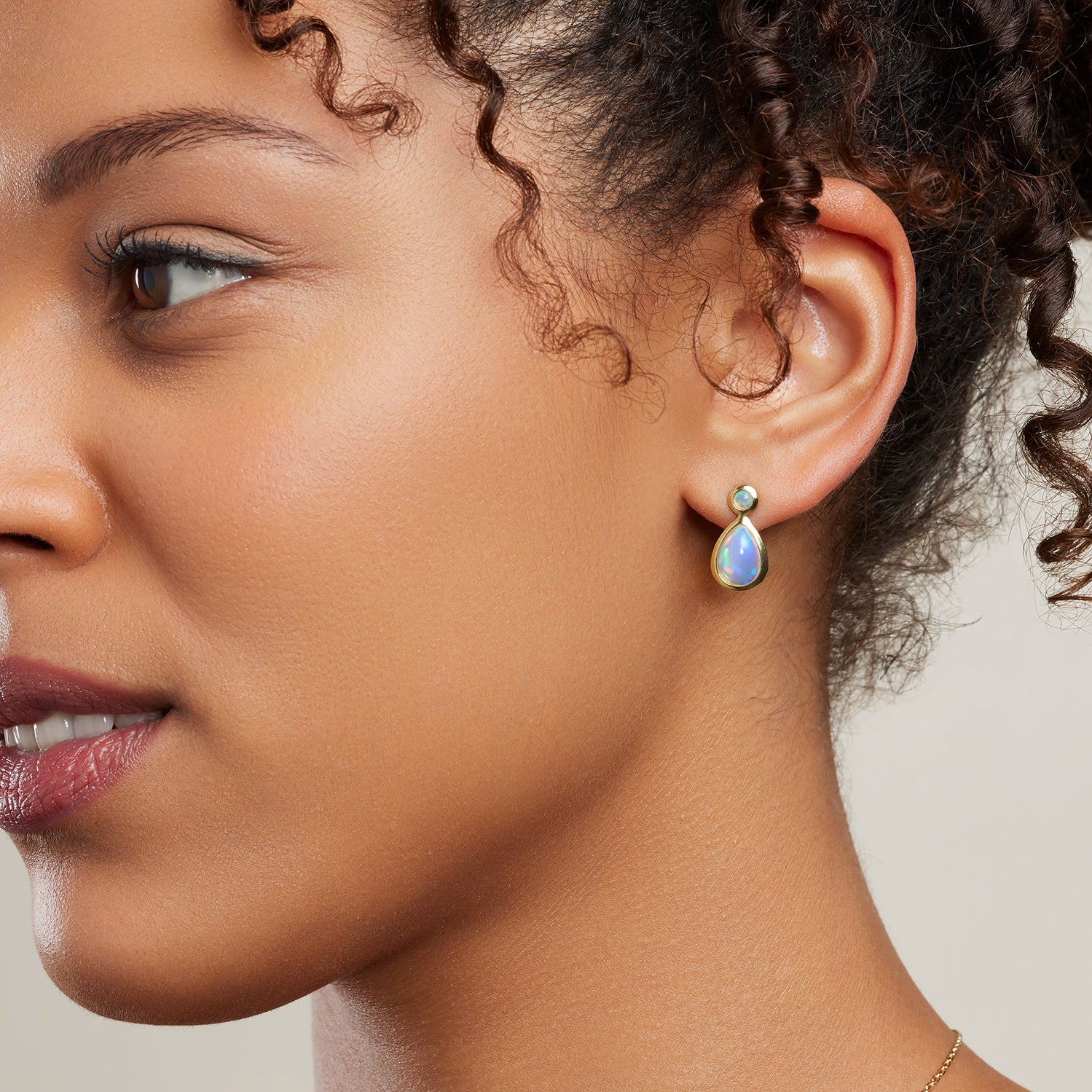 Ausschnitt einer Frau im Profil; sie trägt einen Ohrstecker mit einem kleinen runden, bläulich schimmernden, silbervergoldet eingefassten Opal oben und einem tropfenförmigen größeren Opal unten;
