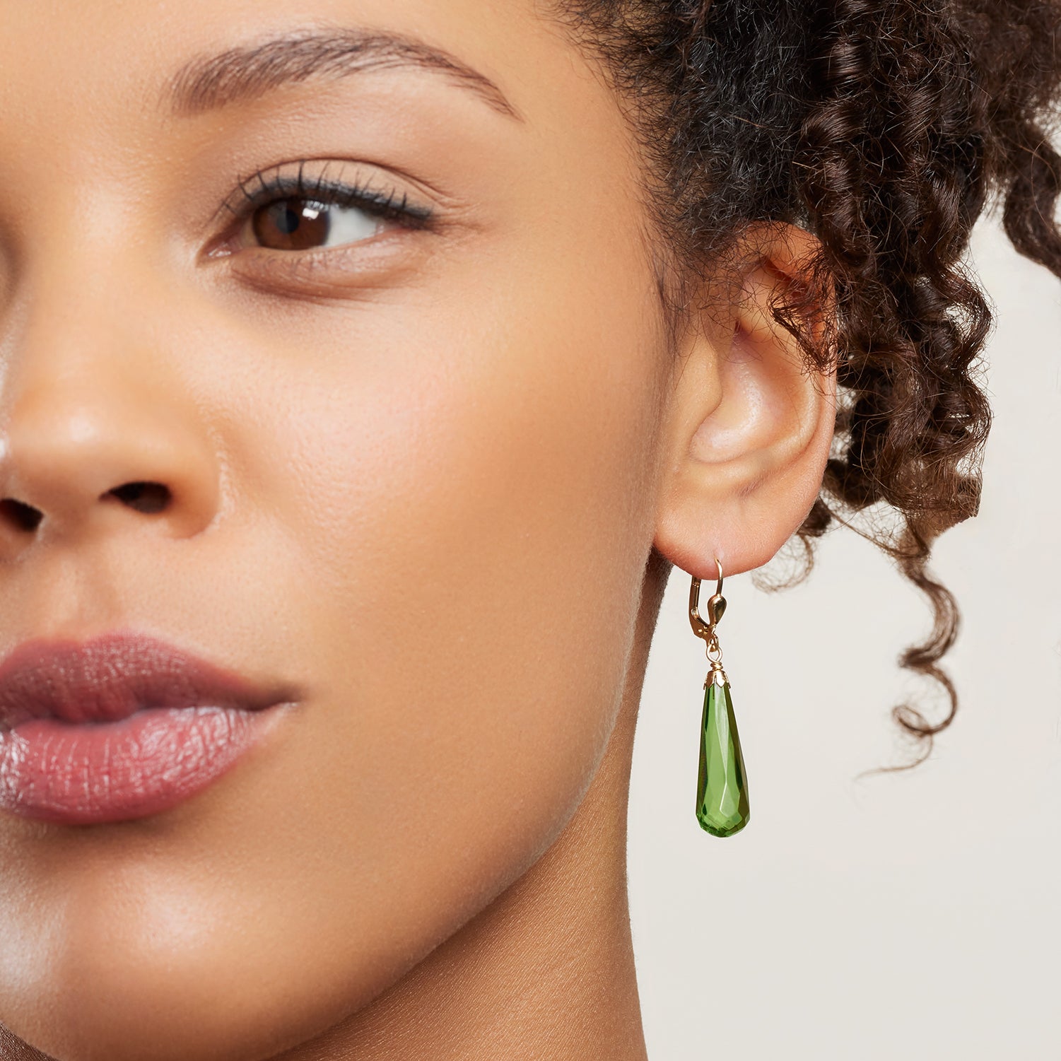Ausschnitt des halben Gesichts einer Frau; sie trägt einen Ohrhänger in Gelbgold mit einem länglichen, grünen Bernstein in Tropfenform;