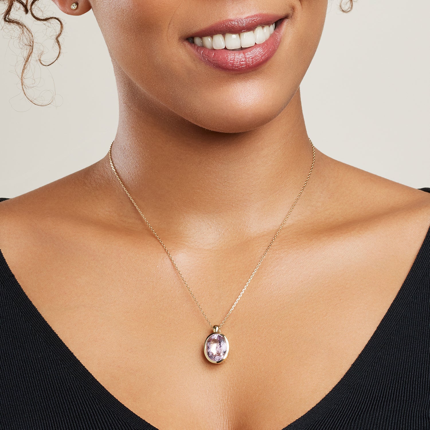 Ausschnitt in Teilen des Gesichts und des Dekolletés einer Frau; sie trägt eine goldene Gliederkette, daran hängt ein goldener Clip-Anhänger mit einem golden eingefassten, ovalen, helllila Stein;