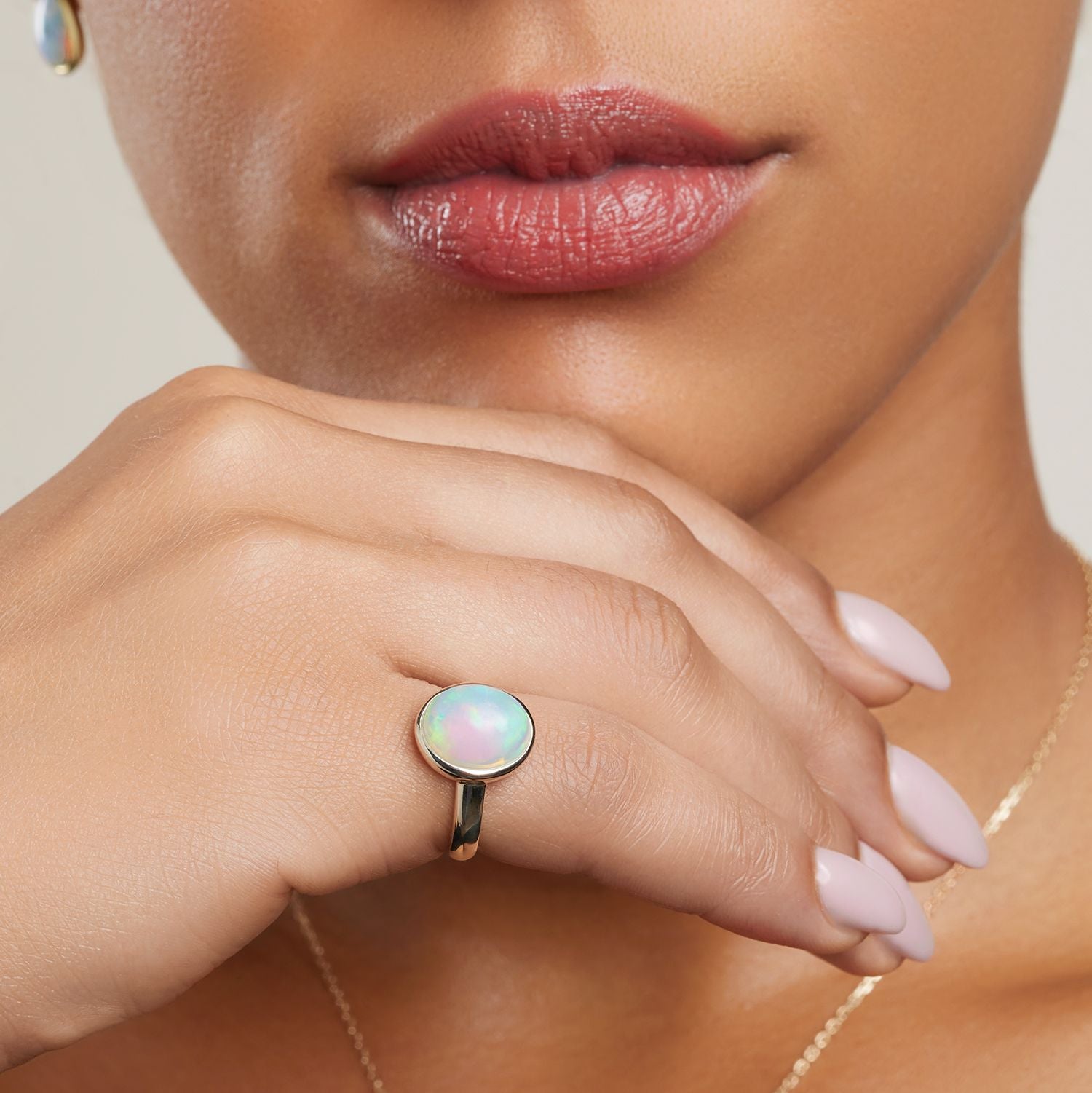 Ausschnitt des Gesichts einer Frau; sie hält ihre Hand an das Kinn und trägt am kleinen Finger einen goldenen Band-Ring mit einem großen, ovalen, bläulich schimmernden Opal;