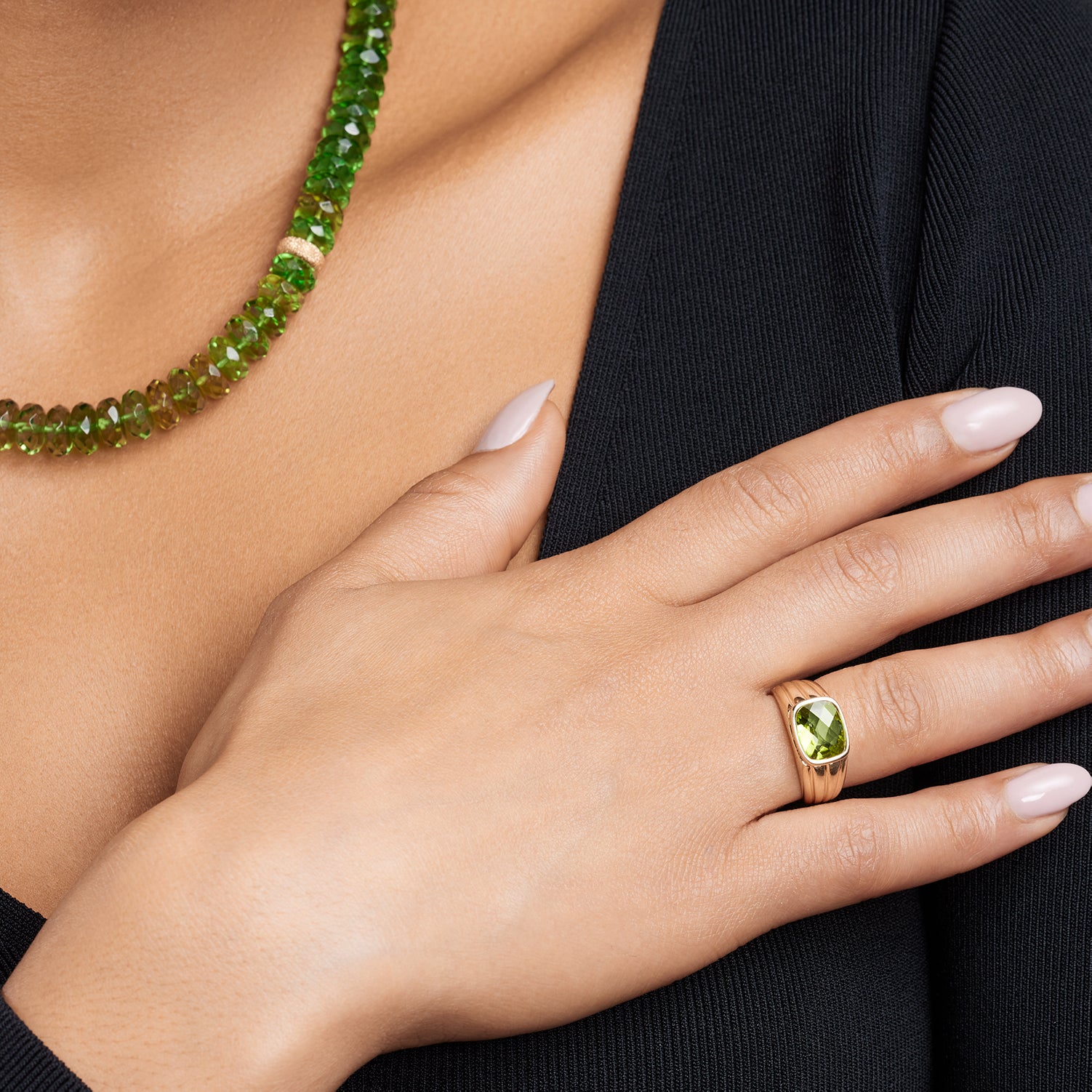 Ausschnitt des Halses und einer Frauenhand, die an die Brust fasst; am Ringfinger ist ein breiter, gerillter Band-Ring in Gelbgold mit einem eingefassten, viereckigen, grünen Stein; am Hals trägt sie ein Collier aus direkt aneinander gereihten, grünen, ovalen Steinen; ein goldener Stein ist eingefügt;