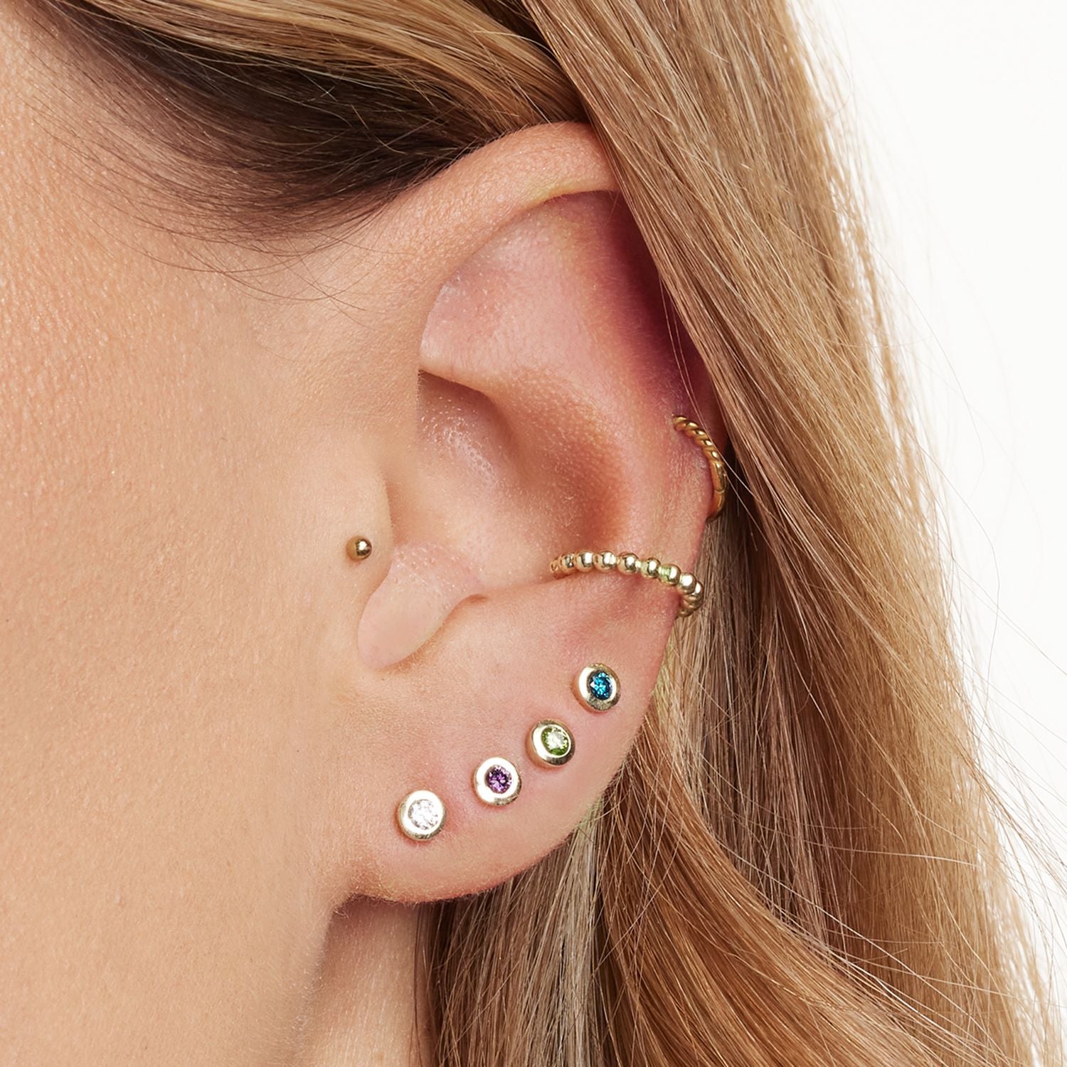 Ausschnitt eines Ohrs mit vier goldenen Ohrstecker mit jeweils einem runden, lila, grünen, blauen und weißen Brillanten in der Mitte; zwei Earcuffs mit goldenen Kugeln und einem kleinen, goldenen Kugel-Ohrstecker;