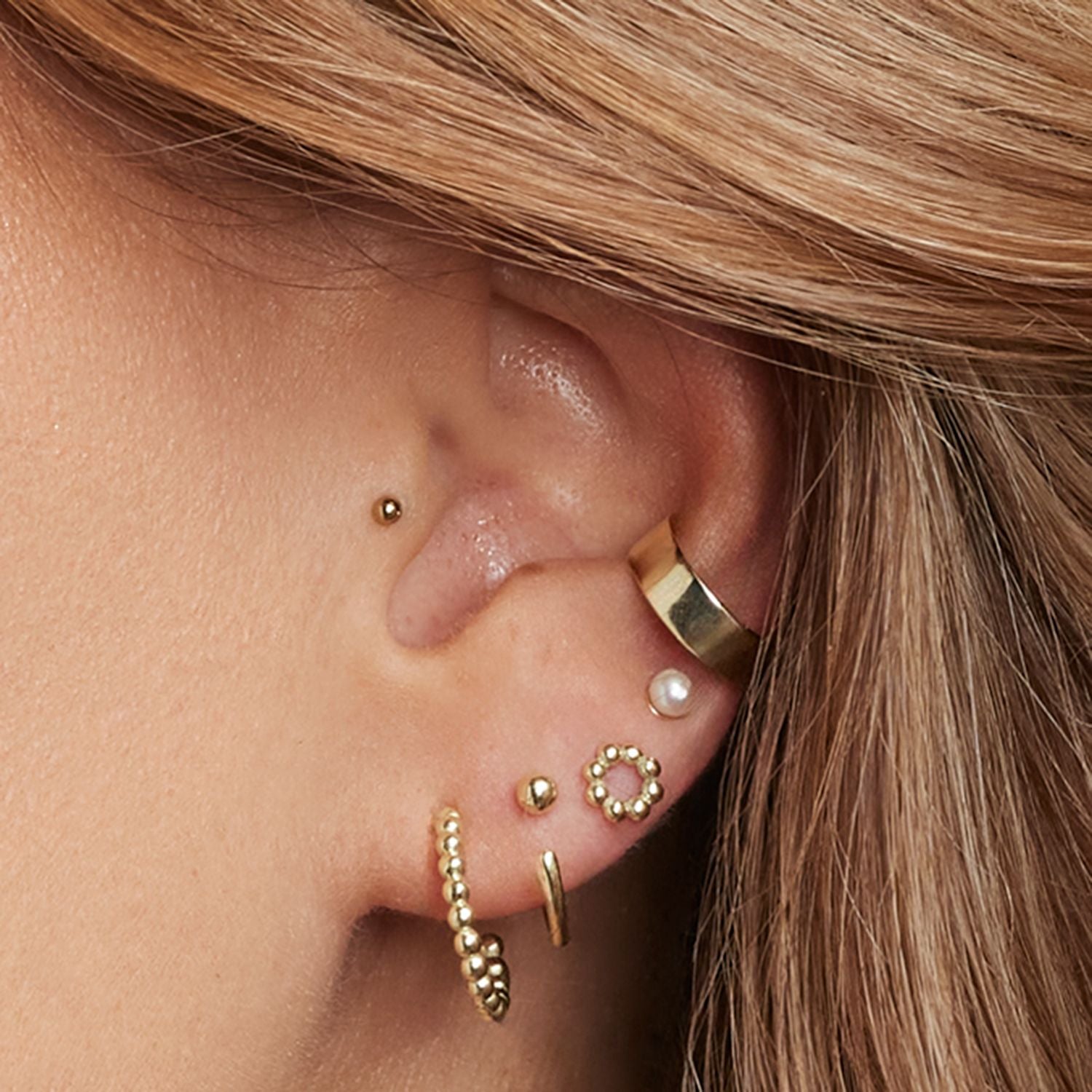 Ausschnitt eines Ohres einer Frau mit einem Ohrsteckern/Earcuffs aus kleinen, goldenen Kugeln, einem Ohrstecker mit einer weißen Perle, einem geraden, goldenen Earcuff, einem Ohrstecker aus kleinen, goldenen Kugeln in Kreisform, einem breiten, goldenen Earcuff;