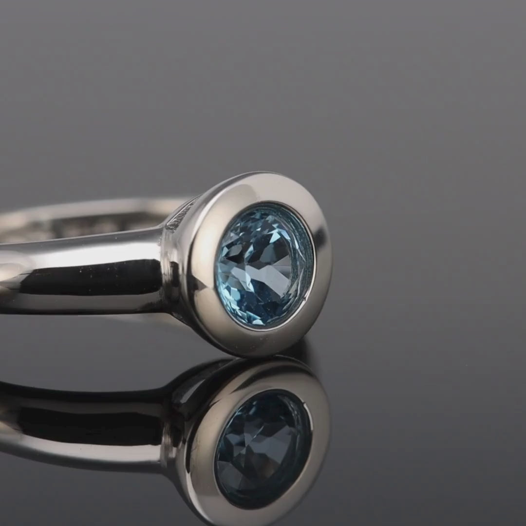 Das Video zeigt einen Band-Ring in Silber mit einem in Silber gefassten, runden, kleinen, blauen Stein; der Ring liegt auf einer spiegelnden Oberfläche;