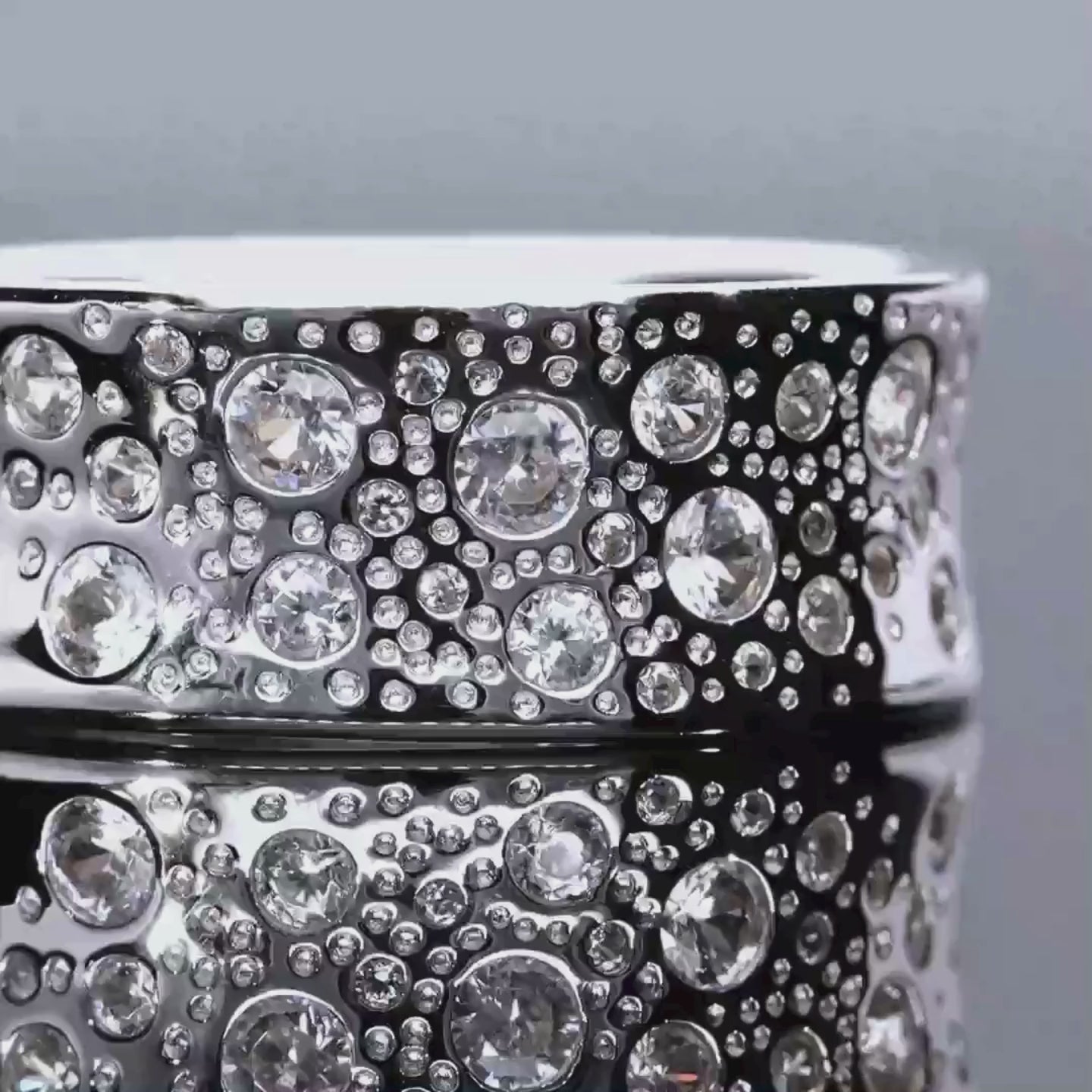 Das Video zeigt einen breiten Band-Ring aus Silber mit vielen eingefassten Zirkonen in unterschiedlichen Größen; der Ring liegt auf einer spiegelnden Oberfläche;