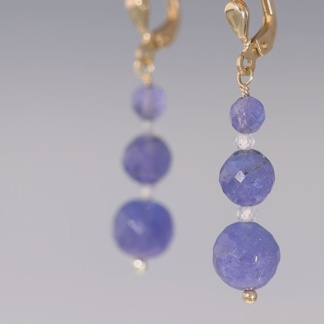 Das Video zeigt Ohrhänger in Gold mit drei runden, jeweils nach unten größer werdenden, blauen Steinen; zwischen den blauen Steinen ist jeweils ein kleiner, weißer, runder Stein; der Ohrhänger schießt unten mit einer goldenen, kleinen Kugel ab;