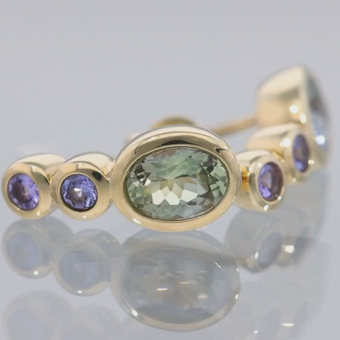 Das Video zeigt einen Ohrstecker in Gelbgold mit einem in Gelbgold gefassten größeren, ovalen grün-blauen Stein und darunter untereinander zwei blau-violette in Gelbgold gefasste, kleinere, runde Steine; der Ohrstecker liegt auf einer spiegelnden Oberfläche;