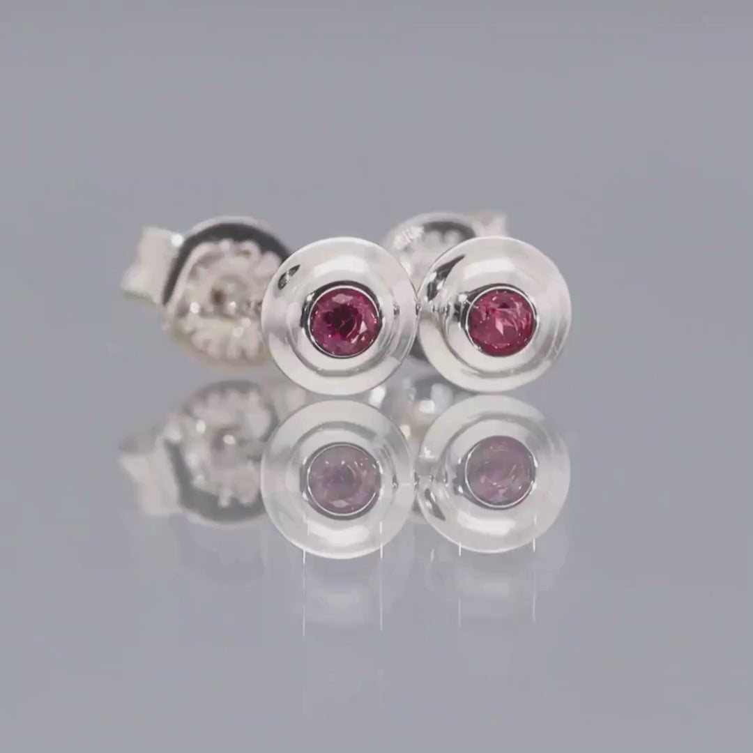 Das Video zeigt zwei runde Ohrstecker (Seitenansicht) in Silber, mit einem lila Stein in der Mitte eingefasst; sie liegen auf einer spiegelnden Oberfläche;
