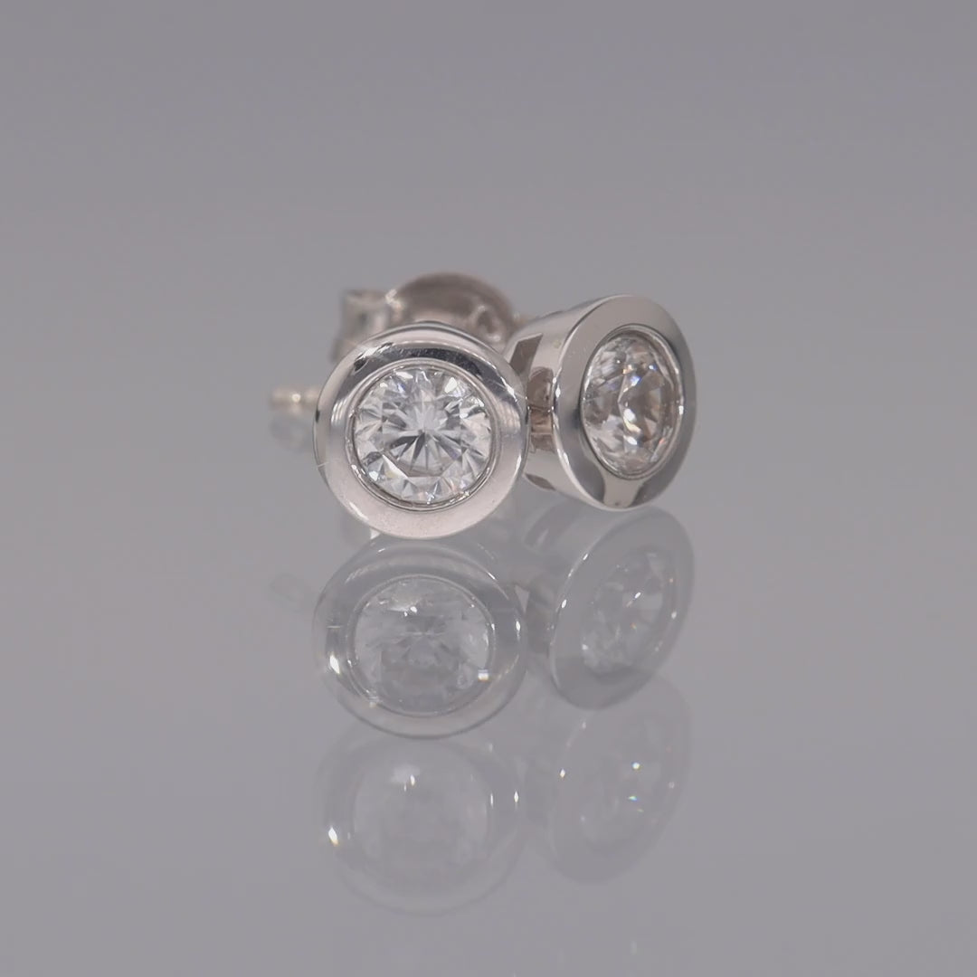 Das Video zweigt zwei runde Ohrstecker in Silber mit einem in Silber gefassten, runden, weißen Zirkon; die Ohrstecker liegen auf einer spiegelnden Oberfläche;