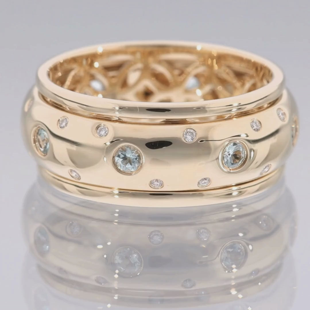 Das Video zeigt einen breiten Band-Ring in Gelbgold mit kleinen vereinzelten Brillanten außen und größeren weißen Steinen in der Mitte; innen ist der Ring nicht glatt sondern offen mit runden Ornamenten; der Ring liegt auf einer spiegelnden Oberfläche;