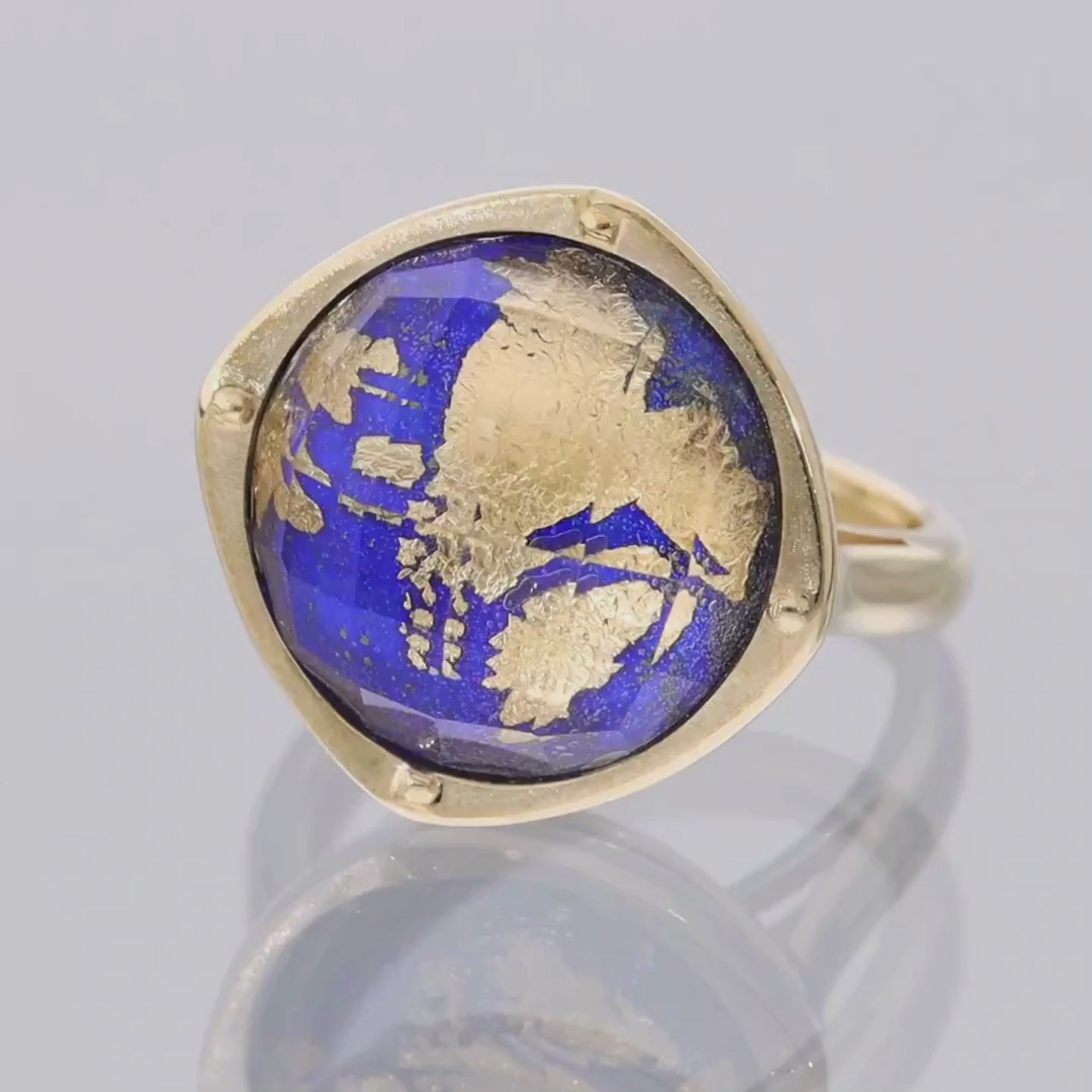 Das Video zeigt einen Ring in Gelbgold mit einem in Gelbgold gefassten, runden, blauen Stein mit Blattgold verziert; der Ring liegt auf einer spiegelnden Oberfläche;