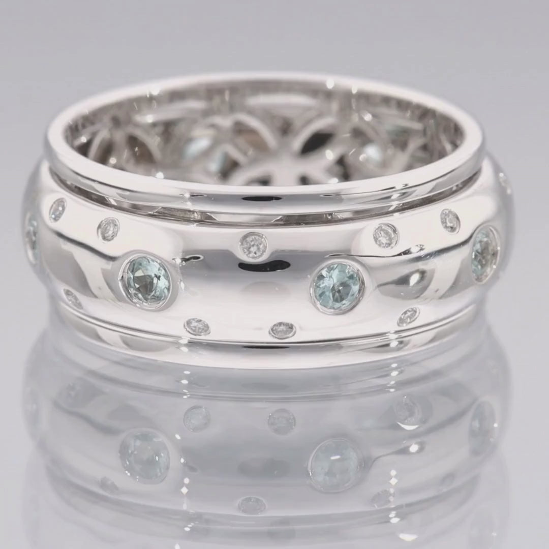 Das Video zeigt einen breiten Band-Ring in Weißgold mit kleinen vereinzelten Brillanten außen und größeren weißen Steinen in der Mitte; innen ist der Ring nicht glatt sondern offen mit runden Ornamenten; der Ring liegt auf einer spiegelnden Oberfläche;