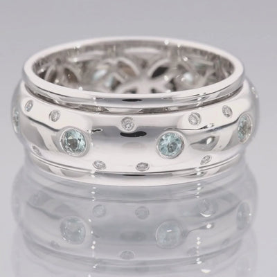 Das Video zeigt einen breiten Band-Ring in Weißgold mit kleinen vereinzelten Brillanten außen und größeren weißen Steinen in der Mitte; innen ist der Ring nicht glatt sondern offen mit runden Ornamenten; der Ring liegt auf einer spiegelnden Oberfläche;
