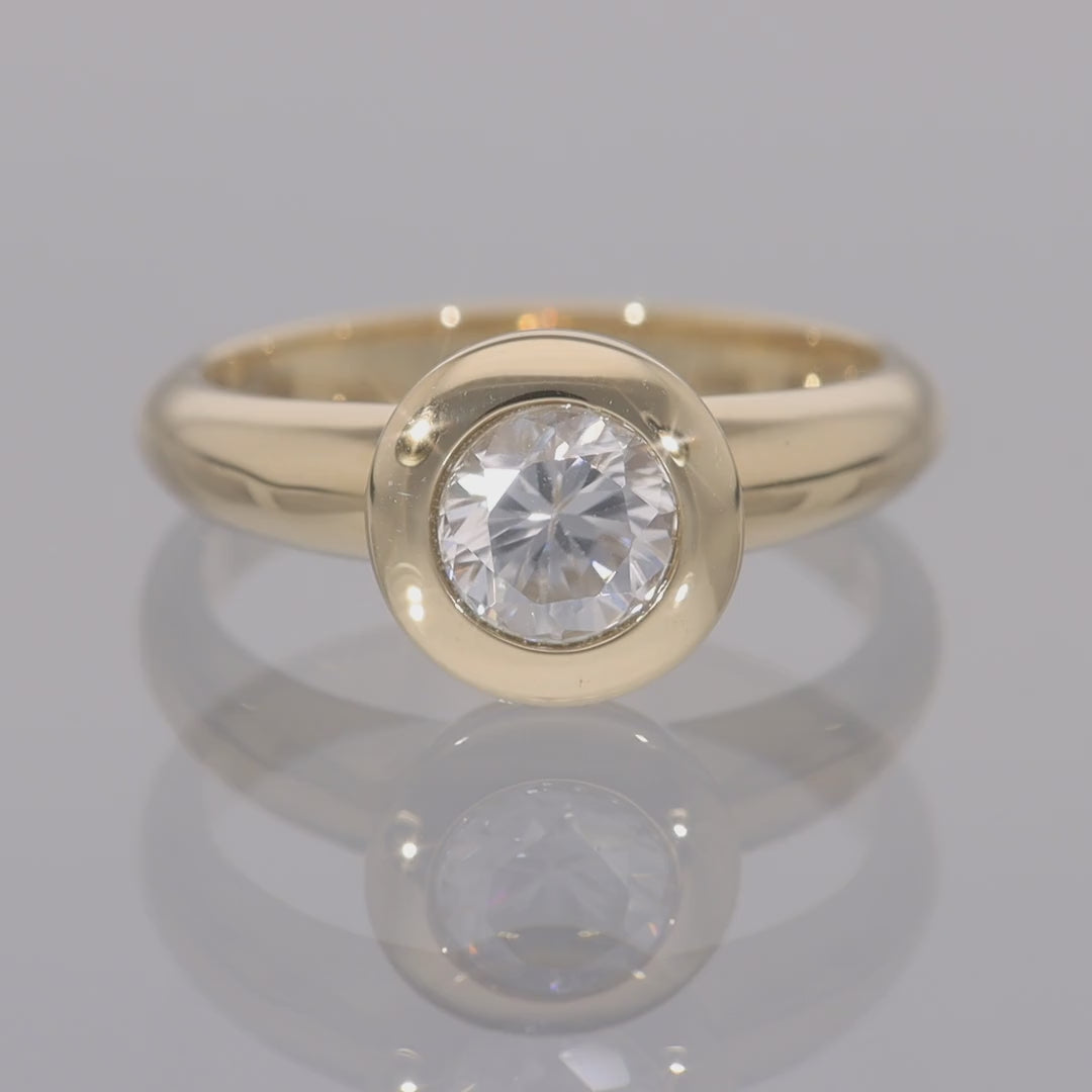 Das Video zeigt einen Band-Ring in Gelbgold mit einem in Gelbgold gefassten, runden, weißen Zirkon; der Ring dreht sich auf einer spiegelnden Oberfläche;