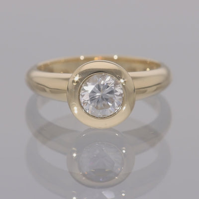 Das Video zeigt einen Band-Ring in Gelbgold mit einem in Gelbgold gefassten, runden, weißen Zirkon; der Ring dreht sich auf einer spiegelnden Oberfläche;