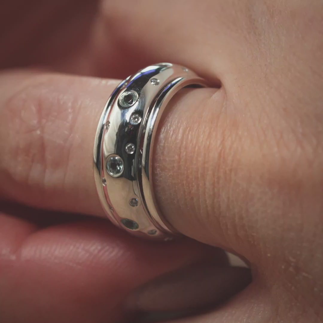 Das Video zeigt den Ausschnitt eines Fingers mit einem breiten Band-Ring in Weißgold mit kleinen vereinzelten Brillanten außen und größeren weißen Steinen in der Mitte; 